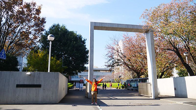 大学の門の前でポーズをとるライターの地主恵亮