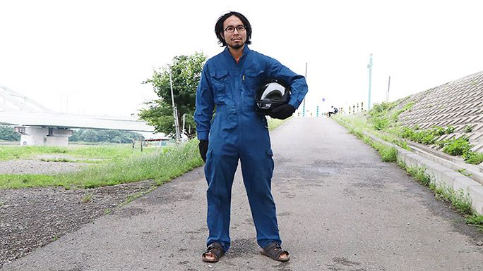 つなぎにヘルメットを抱え、河原でサンダルをはいているライターの地主