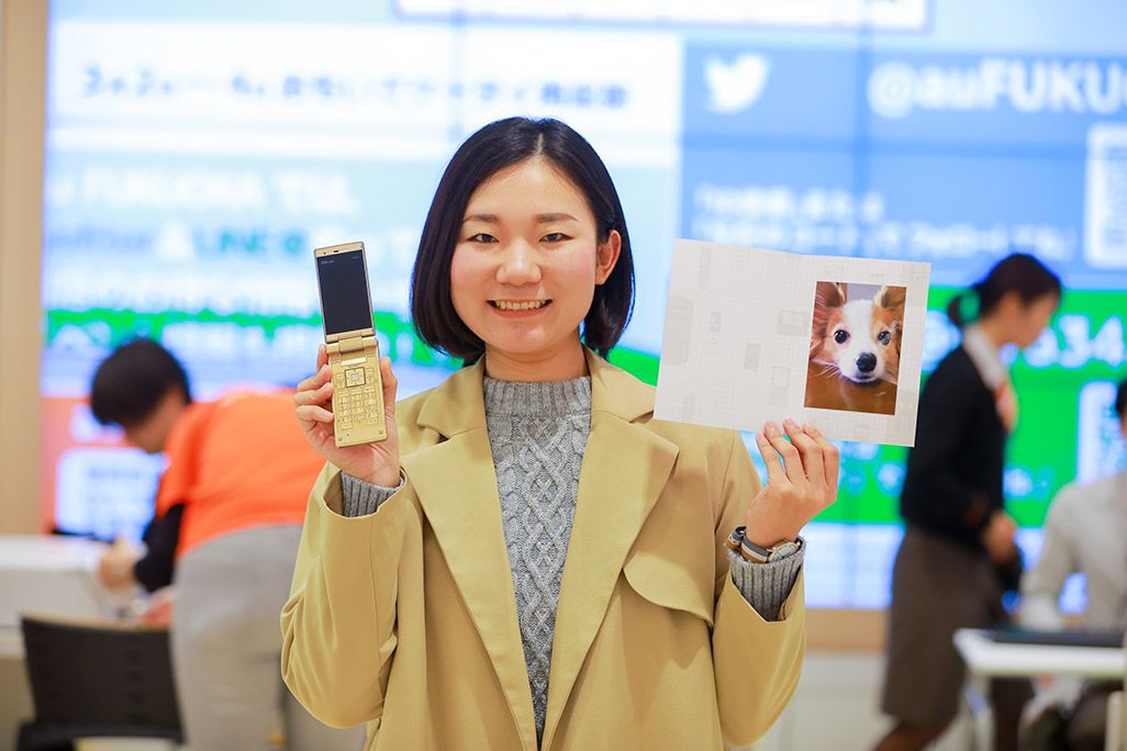au 昔のケータイとプリントアウトされた写真を手に微笑む女性